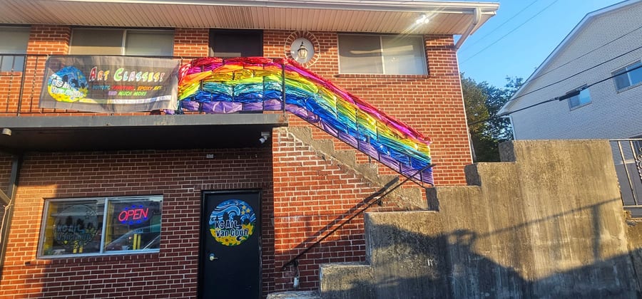 The entrance at 9503 E 63rd Street: a black door with a round KC Art Van Gogh sign, beneath an outdoor staircase decorated with a full rainbow, and an Art Classes banner on the balcony.