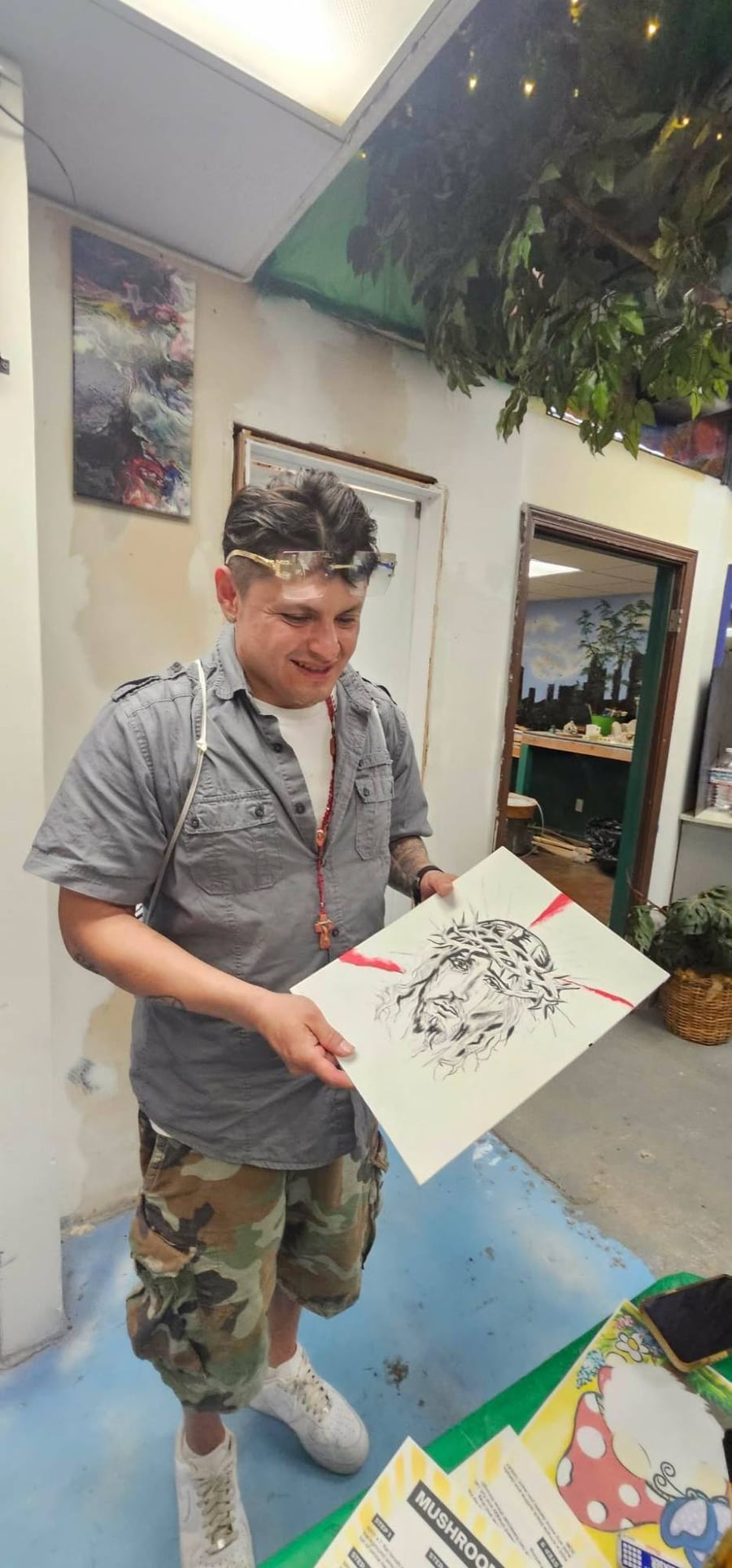 A man in the A.I.M. studio smiling as he holds up his own pen-and-ink drawing of the face of Christ crowned with thorns, with red brushstrokes behind it.