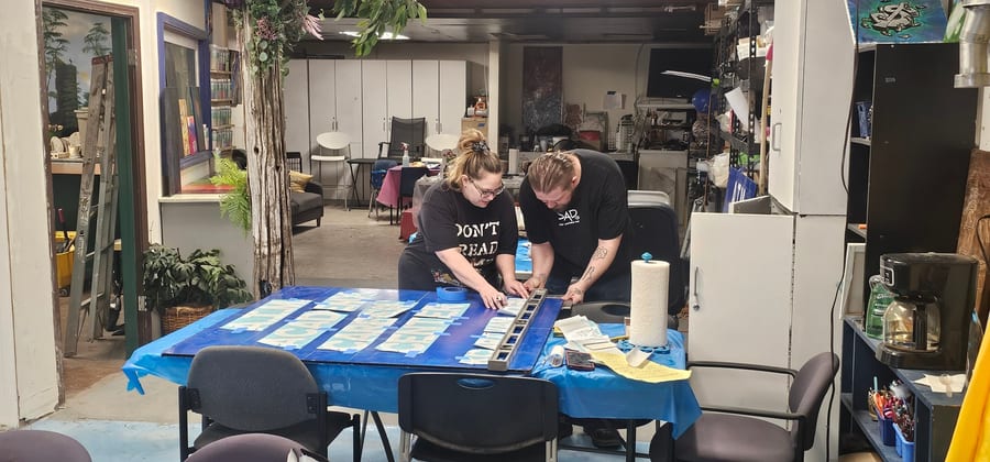 Two people leaning over a table in the A.I.M. studio, aligning white lettering on blue sign panels with a level and masking tape.